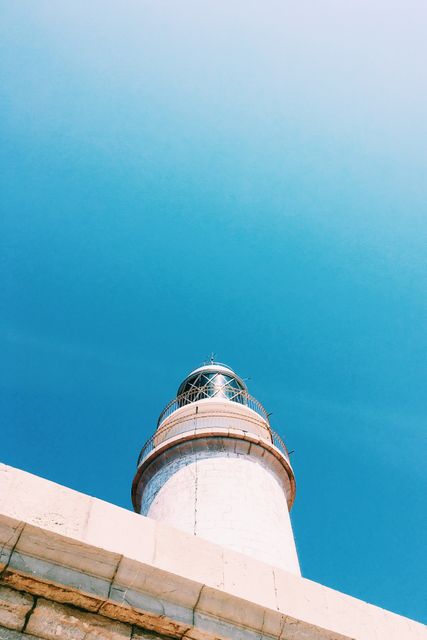 The lighthouse stands tall under a clear blue sky, showcasing its simplistic and minimalistic design. The image can be used in travel blogs, marine navigation articles, inspirational quotes, or decor for coastal-themed spaces. The composition highlights the beauty and utility of the lighthouse with a bright, almost nostalgic atmosphere.