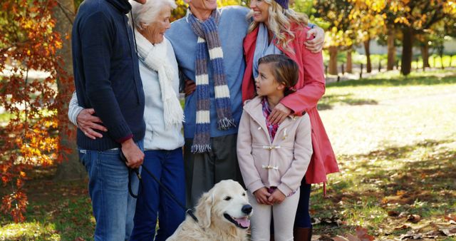 Multi-generational family standing together in park enjoying fall season. Elderly couple with younger adults and child, joined by a golden retriever dog. Leaves changing color in the background, everyone smiling and enjoying time spent outdoors. Ideal for use in family-oriented content, advertisements, promotions about outdoor activities, and content focusing on the joy of family time and seasonal activities.