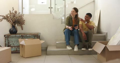 Young couple unpacking moving boxes on stairs in minimalist entryway checking phone