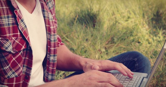 Student is typing on laptop while sitting outdoors on grassy field. Person in casual clothing with plaid shirt enjoys working in nature with portable computer. Ideal for education, nature, work-from-home, or lifestyle topics.