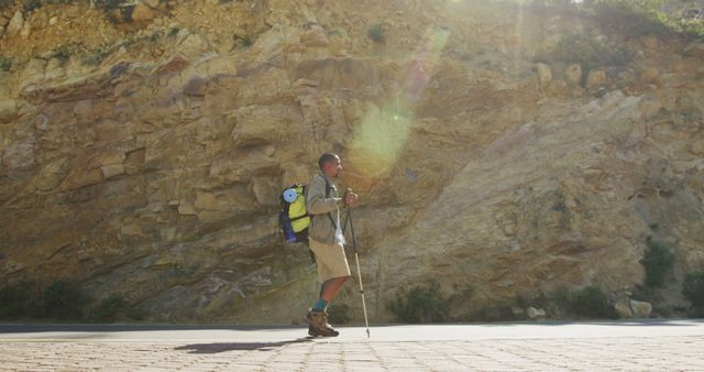 A solo hiker is walking along a path next to a rocky cliffside, equipped with a backpack and hiking gear. The sun is creating a natural lens flare, adding a dramatic effect to the scene. This visual can be used for highlighting outdoor adventures, hiking experiences, travel and exploration themes, promotional materials for outdoor gear, and fitness and lifestyle articles.
