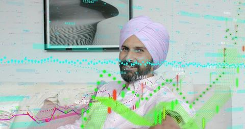Man wearing pink turban analyzing candlestick charts on sofa at home for trading analytics