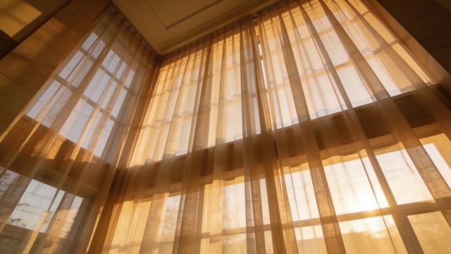 Filtering sheer beige curtains softening golden hour sunlight through tall grid windows