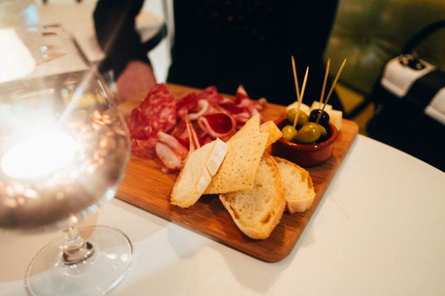 Charcuterie board featuring sliced bread, olives, meat cuts, and cheese on wooden platter. Wine glass and candlelight create a cozy ambience. Perfect for articles or blog posts about dining experiences, gourmet food, restaurant reviews, social gatherings, or evening snacks.