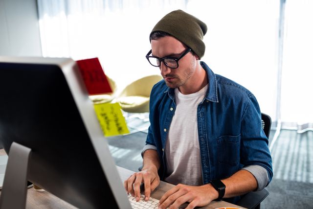 Young hipster man wearing glasses and beanie, working intently at a computer in a modern office. Ideal for use in articles or advertisements about modern work environments, technology, creative industries, and professional lifestyles.