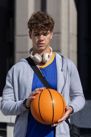 Teen holding basketball wearing headphones in urban plaza, sunlight casting strong shadows