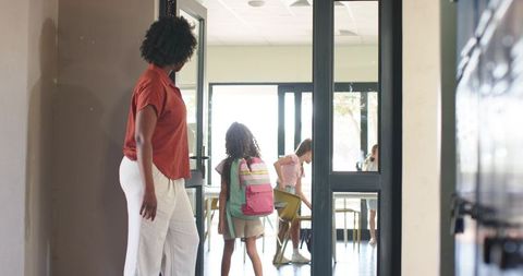 African american teacher guiding students with backpack into bright classroom doorway