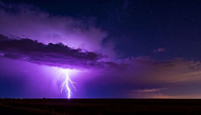 Purple lightning striking open prairie under starry sky with illuminated storm cloud