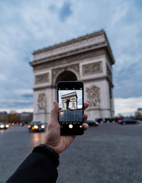 Tourist capturing iconic Arc de Triomphe in Paris with a smartphone, perfect for content about travel, tourism, technology usage, or capturing moments at famous landmarks.