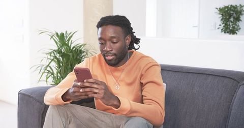 African American Man Sitting on Sofa Using Smartphone in Bright Minimalist Home