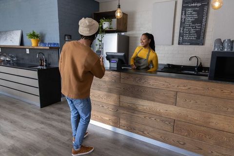 Smiling african american barista serving customer at modern cafe counter using smartphone