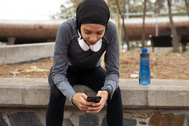 Woman wearing hijab and sportswear taking a break from exercising outdoors in the city, using smartphone with headphones around her neck. Ideal for content related to urban fitness, active lifestyle, technology use in fitness, and promoting diversity in sports.