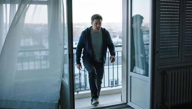 Middle-aged man stepping onto urban balcony wearing hoodie and jeans, reflective mood