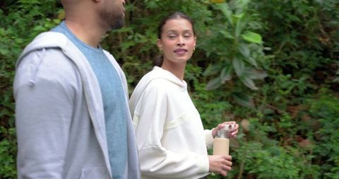 Diverse couple walking wooded trail holding reusable water bottle and wearing hoodies