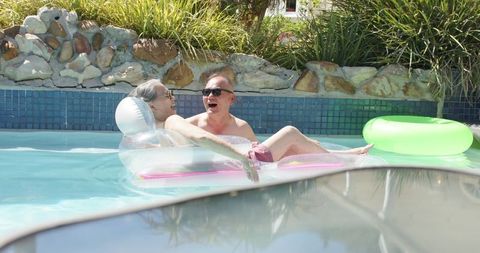 Senior couple relaxing on clear inflatable lounger, floating in sunlit backyard pool