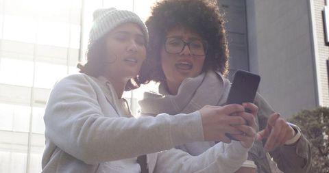 Sharing Moments: Two Young Women Pointing at Smartphone, Laughing and Scrolling in Sunlit City Plaza