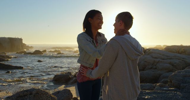 A young Asian woman and a young Caucasian man share a joyful moment on a rocky beach at sunset, with copy space. Their affectionate gaze and smiles suggest a romantic connection as they enjoy the serene coastal environment.