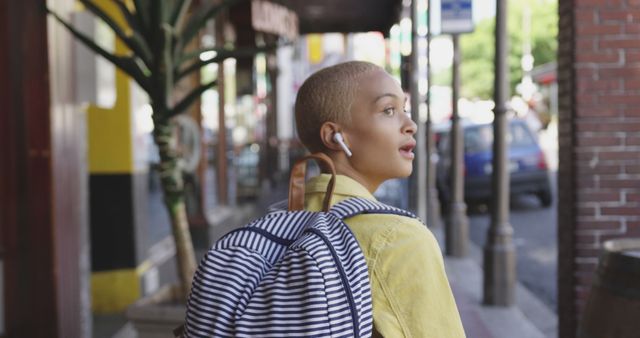 This image portrays a young woman with a shaved head walking in an urban street. She is wearing a striped backpack and listening to something on her wireless earbuds. This visual is perfect for articles or commercials about city lifestyle, travel, student life, technology use, and urban exploration. It can also be used in materials discussing personal style, modern living, summer outings, or street photography.