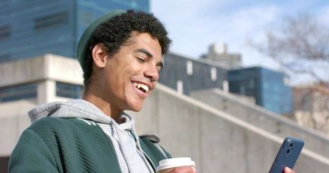 Smiling african american student checking smartphone while holding coffee on urban campus