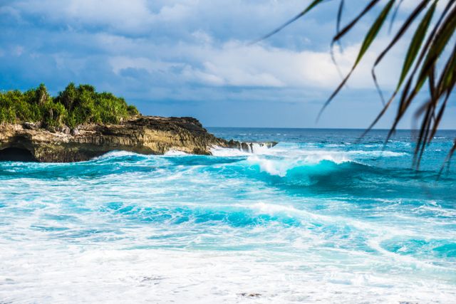 Depicts a vibrant tropical seascape with powerful ocean waves crashing on a rocky shore. Lush greenery is seen atop the rock formations under a cloudy sky, giving a sense of untamed nature and the dynamic beauty of the coast. Ideal for travel brochures, vacation ads, environmental campaigns, and nature-themed projects.