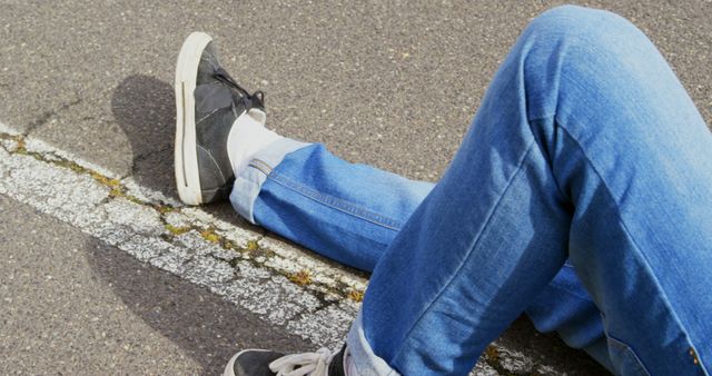 Person sitting on asphalt road wearing blue jeans and black sneakers. Casual and laid-back vibe, making it suitable for use in fashion blogs, lifestyle articles, and social media posts focusing on casual fashion or streetwear.