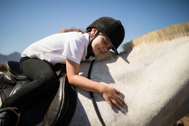 Young girl wearing helmet embracing white horse at ranch on a sunny day. Ideal for use in advertisements, blogs, and articles about horseback riding, children's outdoor activities, animal bonding, and equestrian sports. Perfect for promoting ranches, summer camps, and family-friendly outdoor activities.