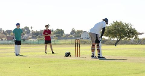 Cricket batsman preparing at crease while teammates watch on sunny suburban field