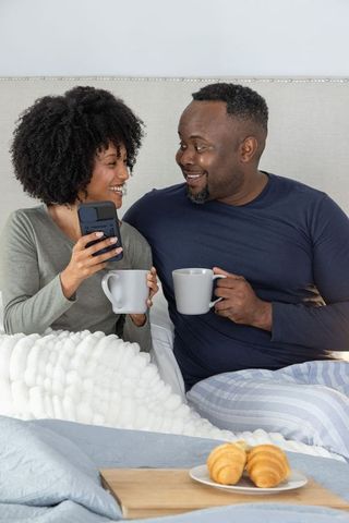African american couple sharing cozy morning in bed with coffee and smartphone