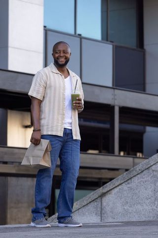 Smiling man holding paper bag and green drink outside contemporary glass office building