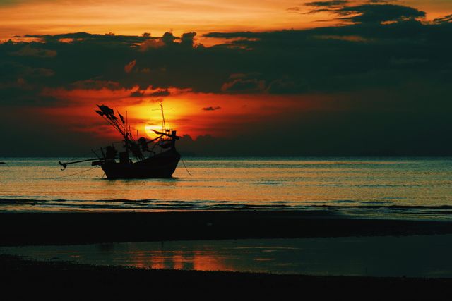 Silhouette of a traditional fishing boat anchored on calm waters with an orange and red sunset in the background, creating a peaceful maritime scene. Sky features dramatic clouds with vibrant colors reflecting on the water. Perfect for travel blogs, nature and landscape photography, or themed decor related to sea and tranquility.