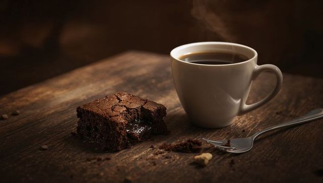 Steaming white ceramic mug beside fudgy chocolate brownie on rustic wooden table, cozy morning