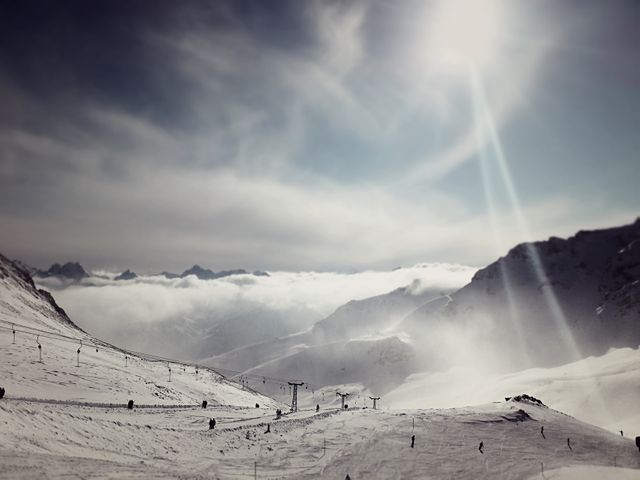 Snow-covered mountains with skiers enjoying winter sport under sunny sky. Ski lift carries people up slopes. Majestic mountains in distance with cloud cover. Perfect for booking a ski trip or promoting winter sports.