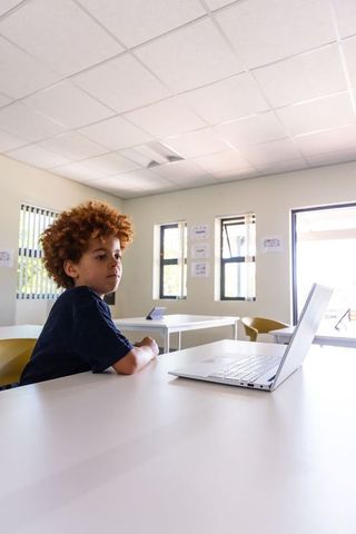 Elementary boy using laptop in bright modern classroom with focused expression