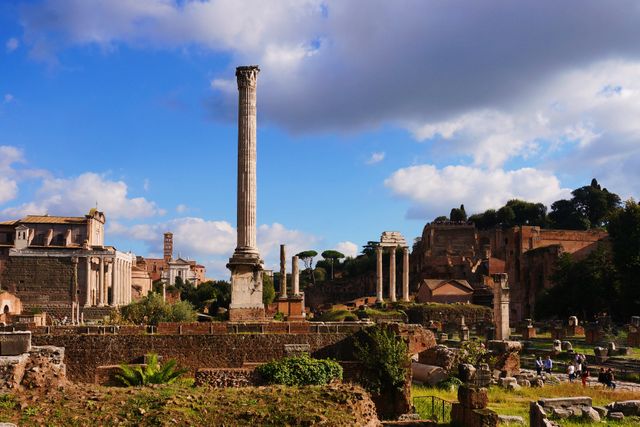 Ancient Roman Forum showing a collection of historic ruins and columns under a blue sky with a few clouds. Useful for travel bloggers, history websites, educational materials, and tourism promotions focusing on historical and archaeological landmarks.