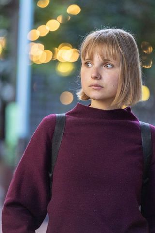 Woman standing on dusk street looking away wearing purple sweater, nose ring, backpack