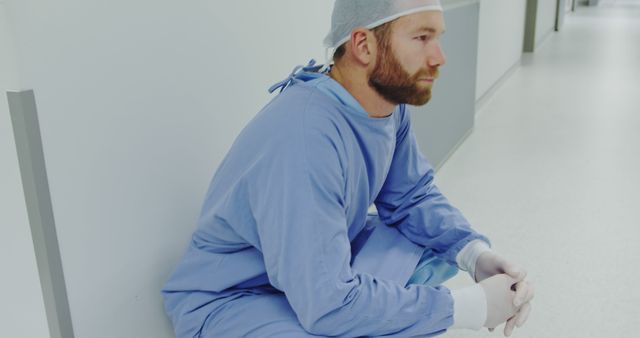 Male doctor in surgical scrubs and cap sitting on hospital floor, appearing exhausted. Ideal for use in articles or posts about healthcare workers’ mental health, the strain of medical professions, the importance of taking breaks, or stories related to the challenges faced by healthcare professionals.