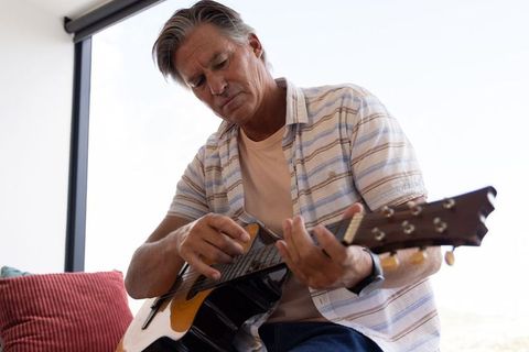 Mature man playing acoustic guitar on couch in sunlit living room, practicing and relaxing