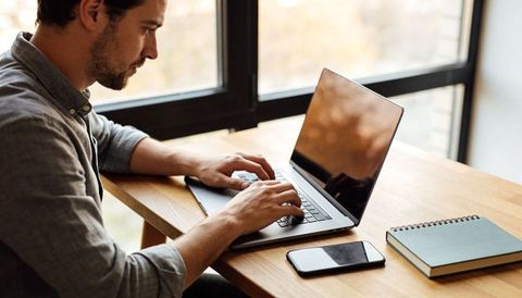 Focused man typing on laptop at wooden desk by window for remote work productivity and focus