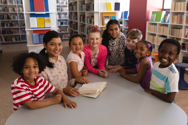 Diverse group of children and female teacher studying together at a table in a school library. They are smiling and looking at the camera, surrounded by bookshelves filled with books. Ideal for educational materials, school brochures, library promotions, and articles on diversity and learning.