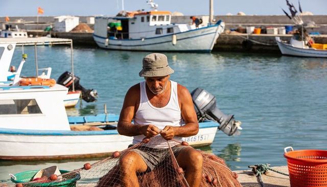 Fisherman mending nets on sunny harbor quay with fishing boats in background