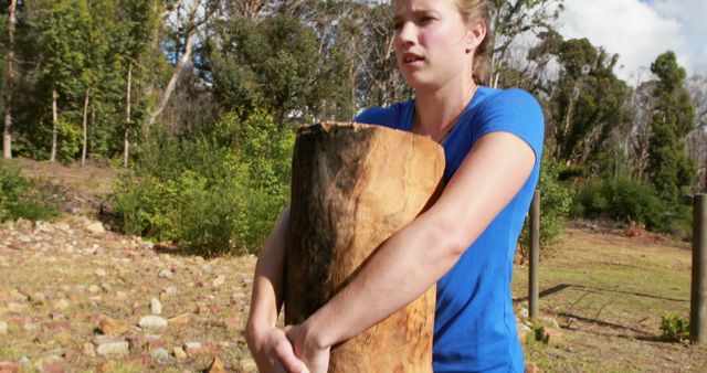 Woman carrying a large wooden log in an outdoor rural area. Suitable for use in articles or publications about rural life, physical labor, outdoor activities, farm life descriptions, and the portrayal of hard work.
