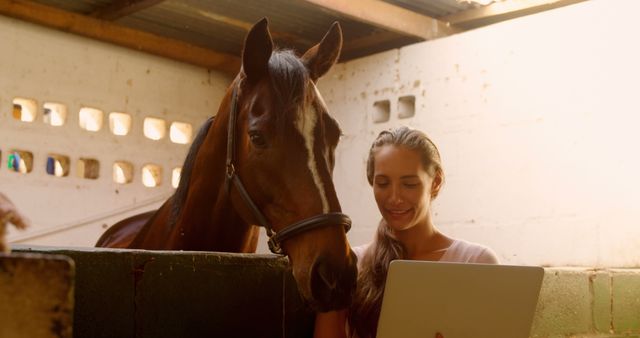 A young Caucasian woman smiles as she uses a laptop near a horse in a stable, with copy space. Her interaction with the animal suggests a blend of technology and equestrian care or training.