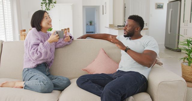 Diverse couple sitting on couch and drinking coffee in living room. Spending quality time at home concept.