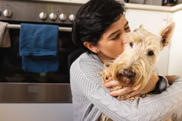Caucasian mid adult lesbian woman with eyes closed embracing and kissing scottish terrier in kitchen. Short hair, unaltered, dog, pet, care, love, togetherness, friendship, lifestyle, home concept.