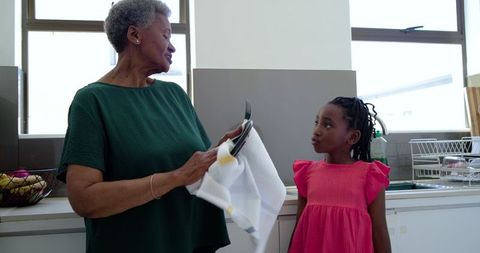 African grandmother drying dishes with granddaughter watching sharing sunlit kitchen moment