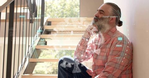 Bearded man sitting on wooden stairs by window touching chin in plaid shirt with holographic overlay