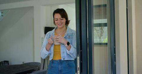 Smiling young woman enjoying morning coffee at sliding glass door in casual denim jacket