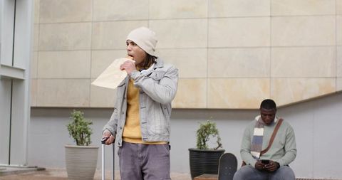 Urban commuters eating sandwich with luggage and skateboard in modern city plaza