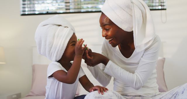 Happy african american mother and daughter wearing towels sitting on bed putting cucumbers on eyes. staying at home in self isolation during quarantine lockdown.