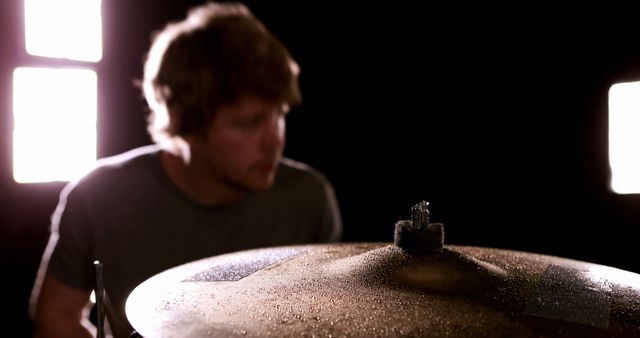 Young male musician playing drums in dimly lit studio with spotlights highlighting the cymbal. Perfect for use in music-related content, promotional materials for bands, educational resources on drumming, artistic photography portfolios, and entertainment industry blogs.